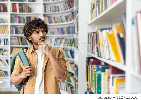 Thoughtful arab customer with backpack on one shoulder holding chin with hand while passing by bookcase. Worried man looking at rows of colourful encyclopaedias and looking for necessary dictionary. Thoughtful arab customer with backpack on one shoulder holding chin with hand while passing by bookcase. Worried man looking at rows of colourful encyclopaedias and looking for necessary dictionary. 112711010