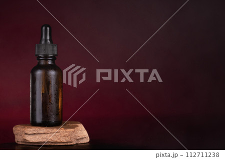 Amber Dropper Bottle on Natural Stone Pedestal with Elegant Maroon Backdrop 112711238