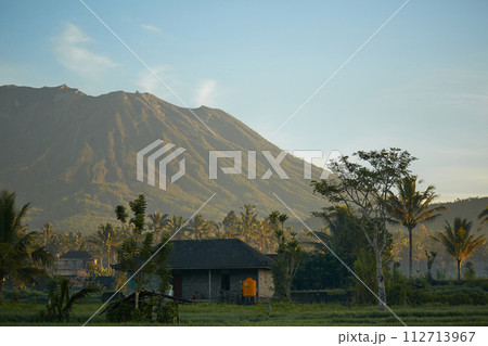 Panorama of Mount Agung and rice fields on the island of Bali. View of the mountain against a background of palm trees and a cornfield. Panorama of Agung volcano covered with clouds on a sunny day. Panorama of Mount Agung and rice fields on the island of Bali. View of the mountain against a background of palm trees and a cornfield. Panorama of Agung volcano covered with clouds on a sunny day. 112713967