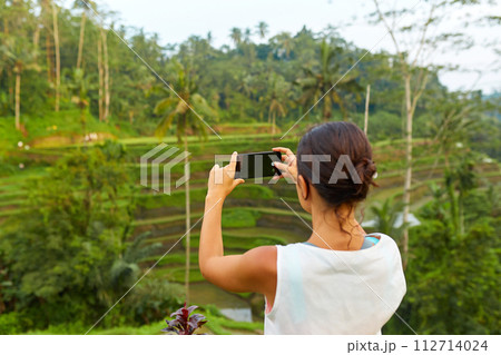 A young slender woman stands with phone on rice terraces on the popular tourist island of Bali. 112714024
