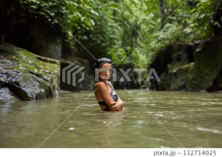A young slender woman in a swimsuit poses by a mountain river in the jungle on the popular island of Bali. A young slender woman in a swimsuit poses by a mountain river in the jungle on the popular island of Bali. 112714025