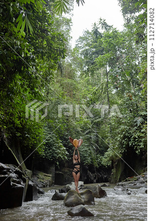A young slender woman in a swimsuit poses with a large dry leaf on a huge boulder covered with moss in the middle of a mountain river in the jungle on the popular island of Bali. 112714082