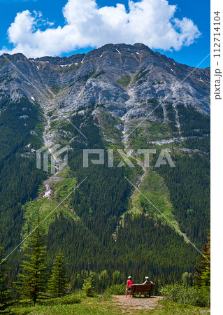 A couple sits on a bench and enjoys the view of a majestic rocky mountain in the Alberta province of Canada. 112714104