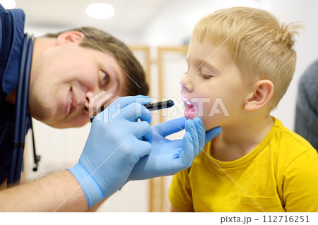 A cute toddler boy with his mother are at an appointment with a pediatrician. The kids ENT doctor examines the throat of a little patient using a flashlight. 112716251