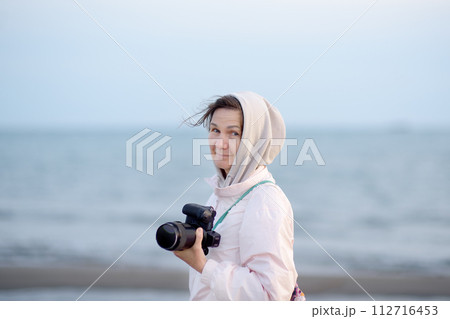 Beautiful elderly lady walking along the sea shore. Portrait of a senior woman photographer with camera. Single travel Beautiful elderly lady walking along the sea shore. Portrait of a senior woman photographer with camera. Single travel 112716453