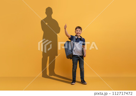 A young boy with a backpack gives a thumbs up in front of a yellow background 112717472