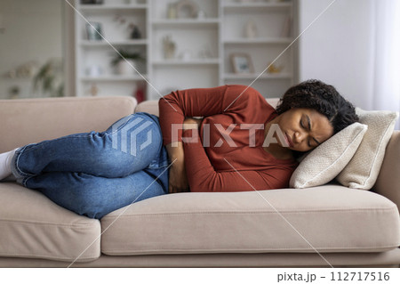 Young black woman clutching her abdomen in discomfort at home 112717516