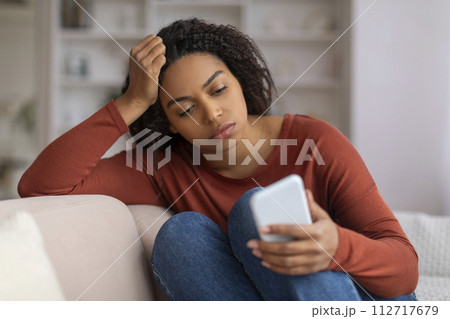 Young black woman feeling concerned while looking at her smartphone at home Young black woman feeling concerned while looking at her smartphone at home 112717679