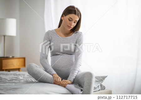 Young pregnant woman in casual wear massaging her swollen ankle at home 112717715