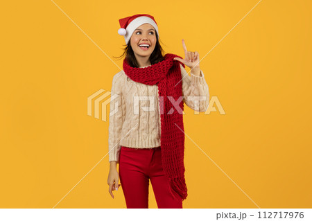 woman in Santa hat and scarf pointing upward, yellow backdrop woman in Santa hat and scarf pointing upward, yellow backdrop 112717976