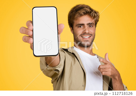 European man promotes mobile app, pointing at phone on yellow background 112718126