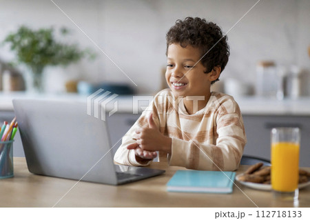 Happy black boy interacting with laptop while sitting at table in kitchen Happy black boy interacting with laptop while sitting at table in kitchen 112718133