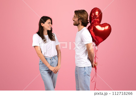 A happy couple stands facing each other with a playful expression, holding shiny red heart-shaped balloons A happy couple stands facing each other with a playful expression, holding shiny red heart-shaped balloons 112718266