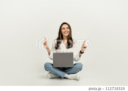 Happy woman sitting cross-legged on the floor with a laptop, pointing upwards 112718320