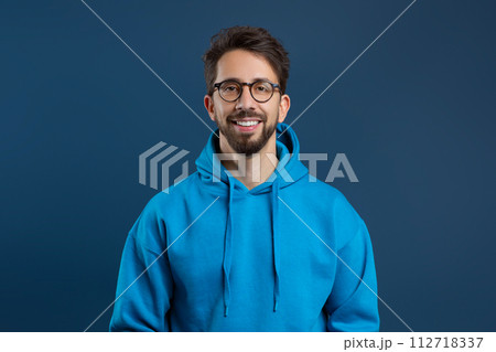 Cheerful young man with beard and glasses standing against dark blue background 112718337