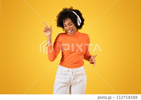 Joyful young woman with curly afro hair wearing headphones dances and points upwards 112718388