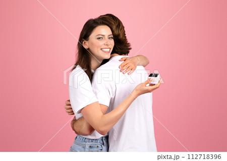 Delighted woman with a bright smile hugging a man while sneakily holding an engagement ring box behind his back Delighted woman with a bright smile hugging a man while sneakily holding an engagement ring box behind his back 112718396