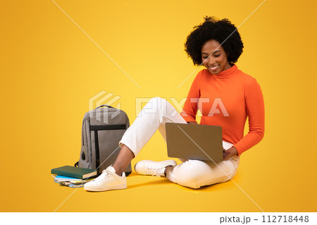 Engaged African American student with curly hair using a laptop, sitting cross-legged on the floor 112718448