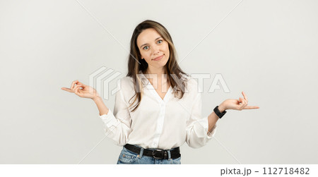 A young woman with long brown hair, wearing a white blouse and jeans, stands with a playful shrug A young woman with long brown hair, wearing a white blouse and jeans, stands with a playful shrug 112718482