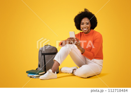 A content young woman with a curly afro sits on the floor with her smartphone 112718484