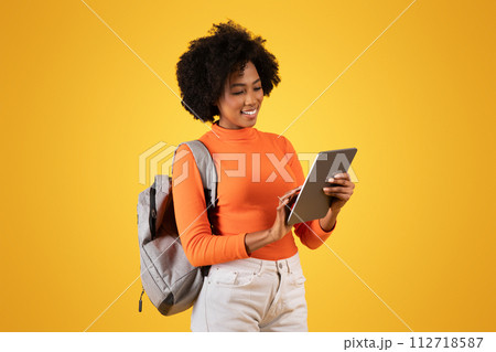 Engrossed African American woman with a delightful smile using a tablet, carrying a backpack 112718587