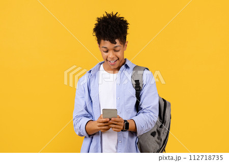 Happy black male student with smartphone and backpack against yellow background Happy black male student with smartphone and backpack against yellow background 112718735
