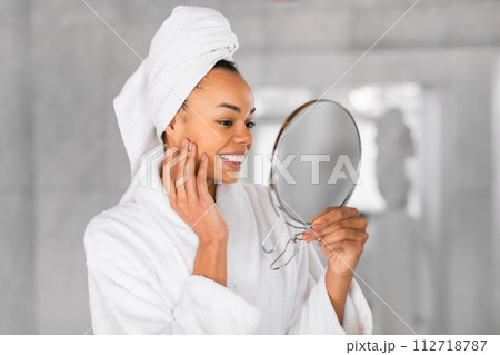 Pretty black woman holds mirror gently touching face in bathroom 112718787