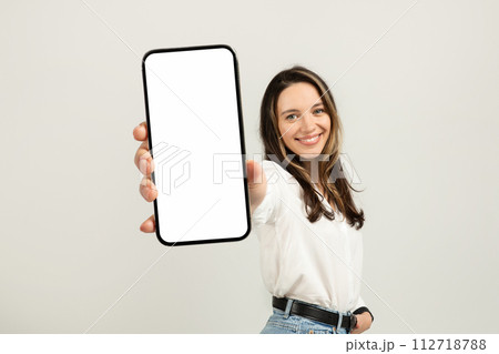 Smiling young brunette woman in a white blouse presenting a smartphone with a blank screen 112718788