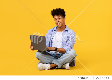 Smiling black student guy with laptop sitting cross-legged on yellow background Smiling black student guy with laptop sitting cross-legged on yellow background 112718833