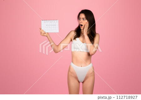 Shocked young woman in white underwear holding a calendar with a surprised expression 112718927