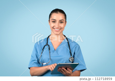 Attentive woman nurse in scrubs with tablet smiling at camera 112718959