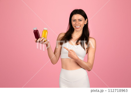 A cheerful young woman in a white sports bra and high-waisted leggings holding two bottles of juice 112718963