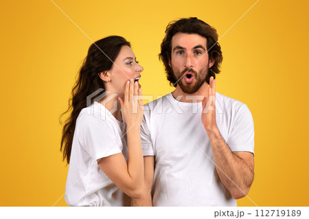 Surprised bearded man with wide eyes and an open mouth holds his hands up while a woman whispers into his ear Surprised bearded man with wide eyes and an open mouth holds his hands up while a woman whispers into his ear 112719189
