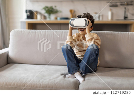 Excited black boy wearing virtual reality headset sitting on couch at home Excited black boy wearing virtual reality headset sitting on couch at home 112719280