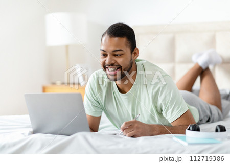Happy african american guy watching webinar, bedroom interior Happy african american guy watching webinar, bedroom interior 112719386