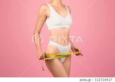 A fit woman in white underwear measures her waist with a yellow tape measure 112719417