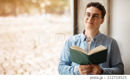 A contented young man with curly hair and round glasses is engrossed in reading a book A contented young man with curly hair and round glasses is engrossed in reading a book 112719525