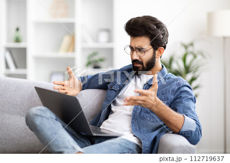 Confused Young Indian Guy Suffering Problems While Using Laptop At Home Confused Young Indian Guy Suffering Problems While Using Laptop At Home 112719637