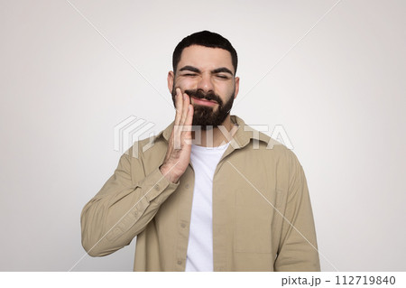Man with a beard winces in discomfort, touching his cheek with a hand 112719840