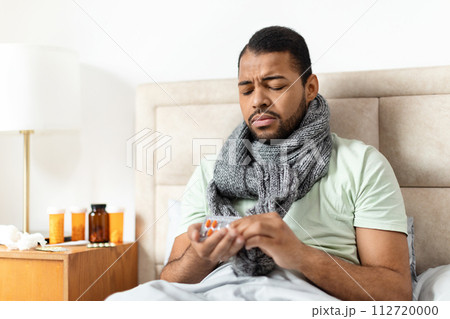 Sick african american man sitting in bed, taking pills Sick african american man sitting in bed, taking pills 112720000