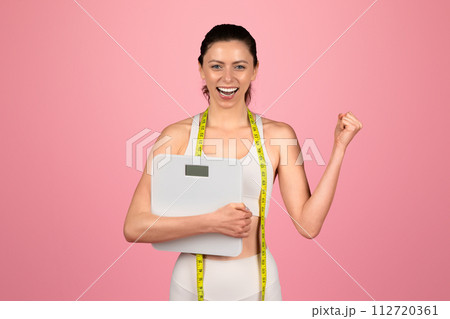 Exultant young woman celebrating her weight loss success, holding a scale and wearing a measuring tape 112720361