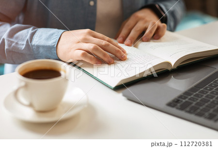 Close-up of a person's hands reading a book with a cup of coffee on the side and a laptop in the background 112720381