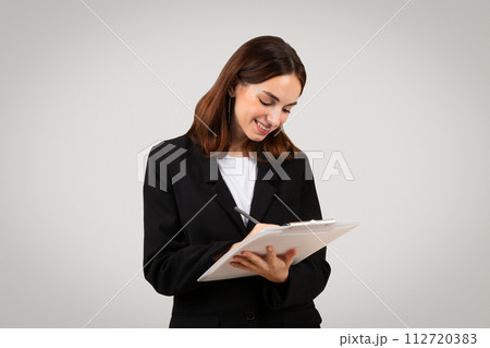 Smiling businesswoman joyfully jotting down notes on a clipboard, displaying a moment of productivity Smiling businesswoman joyfully jotting down notes on a clipboard, displaying a moment of productivity 112720383