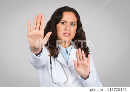 Young lady doctor making stop gesture, keeping hands to defend, protect herself, standing on gray background Young lady doctor making stop gesture, keeping hands to defend, protect herself, standing on gray background 112720393