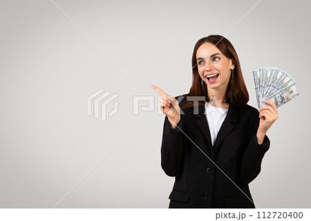 Excited businesswoman in a black suit holding a fan of dollar bills and pointing to the side Excited businesswoman in a black suit holding a fan of dollar bills and pointing to the side 112720400