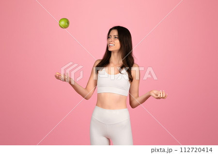 Playful young woman in white fitness attire joyfully tossing a green apple into the air Playful young woman in white fitness attire joyfully tossing a green apple into the air 112720414