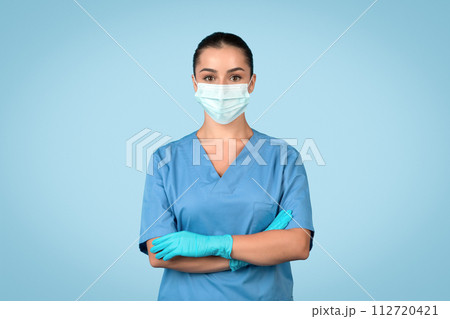 Young woman nurse in scrubs with mask and gloves on blue backdrop 112720421