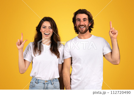 A joyful woman and a man with beards point upwards with bright smiles, wearing white t-shirts A joyful woman and a man with beards point upwards with bright smiles, wearing white t-shirts 112720490