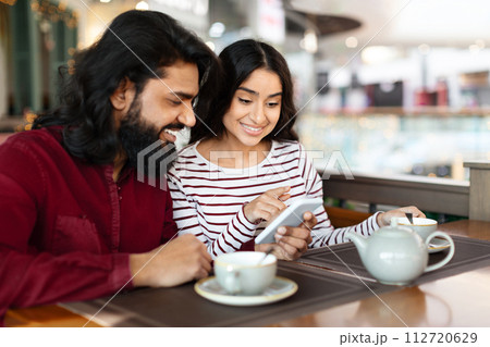 Happy young indian woman show phone to boyfriend, cafe interior Happy young indian woman show phone to boyfriend, cafe interior 112720629