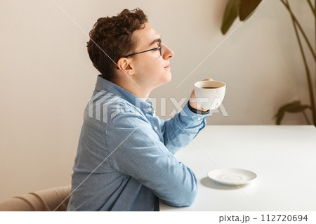 Relaxed young man with curly hair and glasses enjoying a moment of reflection with a hot cup of coffee 112720694